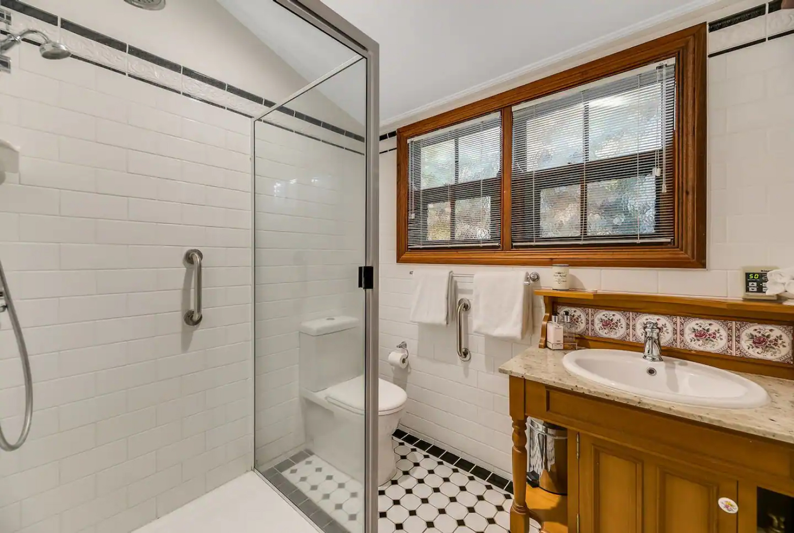 This image shows a bathroom with a glass-enclosed shower, white subway tiles, and a black and white checkered floor. There is a wooden vanity with a sink and a mirror above it. The window has blinds, and there are towels hanging on a rail.