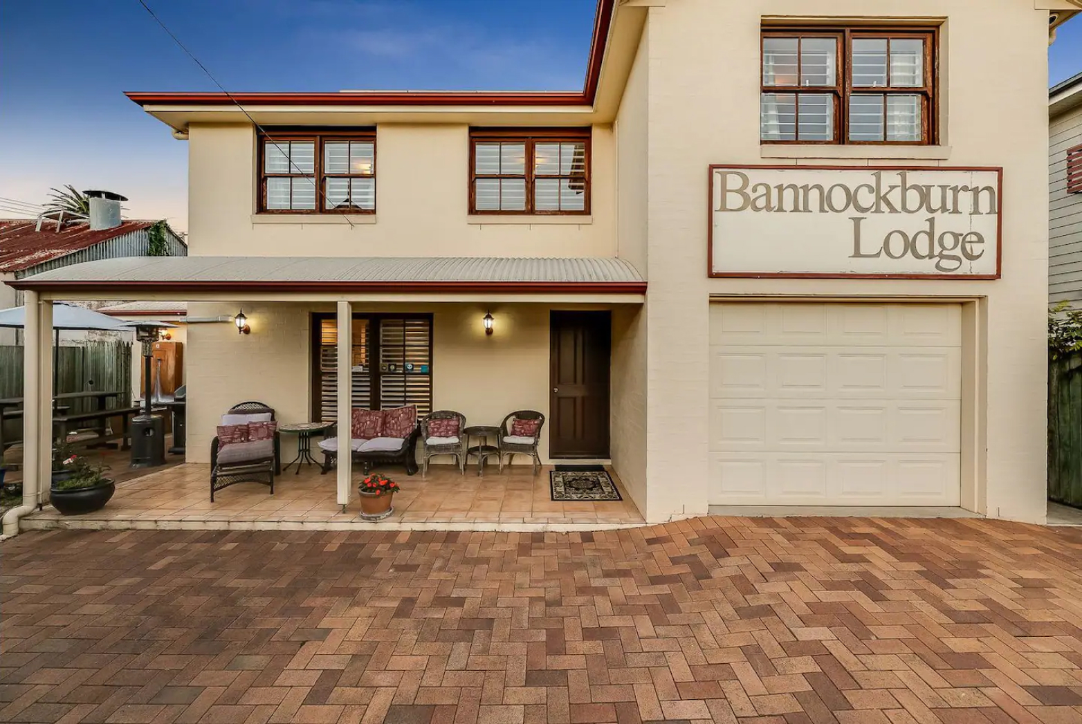 The image shows a two-story building with a sign that reads "Bannockburn Lodge." The exterior is cream-colored with brown trim. There is a covered patio area with outdoor furniture, including chairs and a table. The ground is paved with bricks