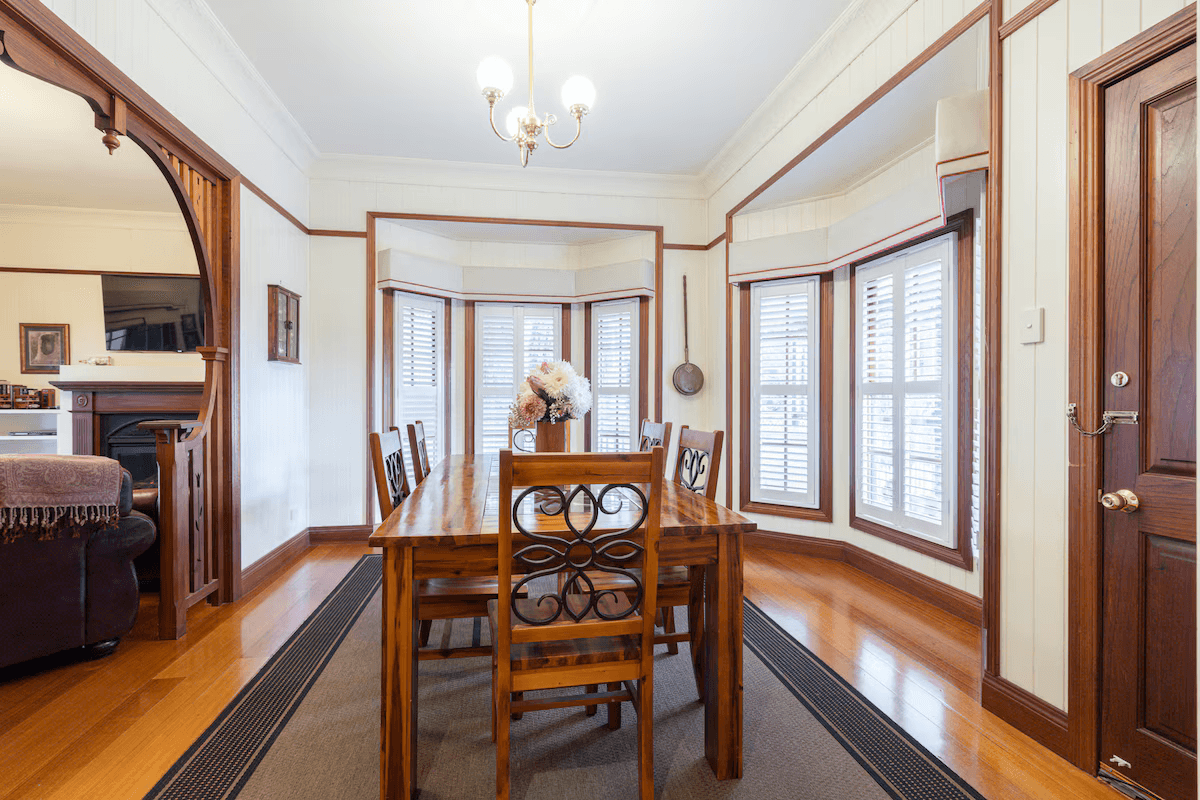 Elegant dining room with wooden table and chairs, polished hardwood floors, bay windows with white shutters, and soft natural light