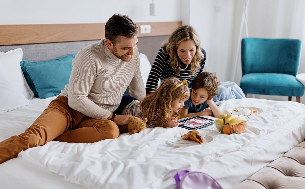 A happy family with two children relaxing on a bed while watching a tablet and enjoying breakfast together